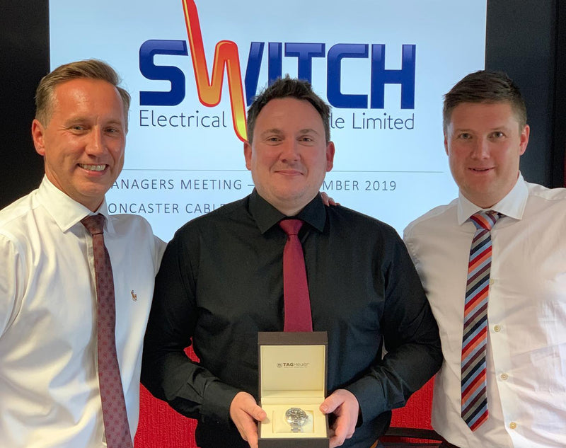Three men standing together, with one holding an award, in front of a Switch Electrical Limited sign.